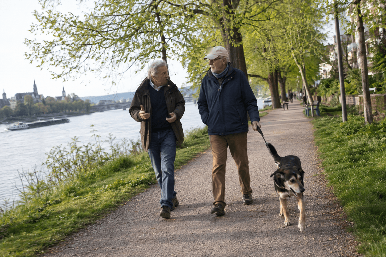Zwei ältere Männer unterhalten sich beim Spaziergang mit Hund auf einem Uferweg im Frühling