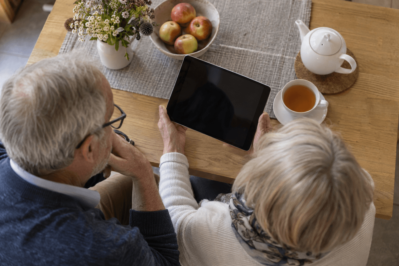 Älteres Paar blickt am Esstisch gemeinsam auf ein Tablet, daneben Tee und eine Schale Äpfel