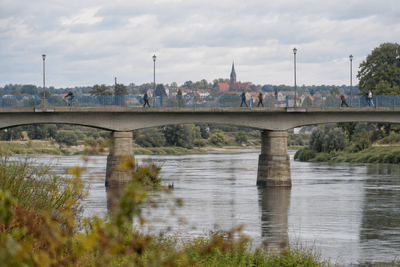 Fußgänger und ein Radfahrer überqueren an einem bewölkten Tag eine Brücke über einen breiten Fluss, dahinter eine Kleinstadt mit Kirchturm