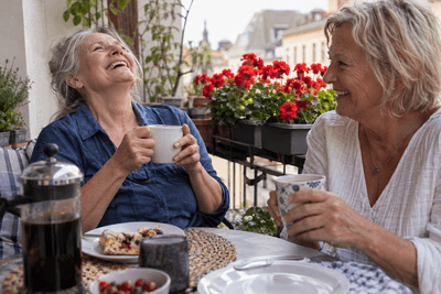 Zwei Frauen um die sechzig lachen beim Kaffeetrinken auf einem Balkon mit roten Geranien